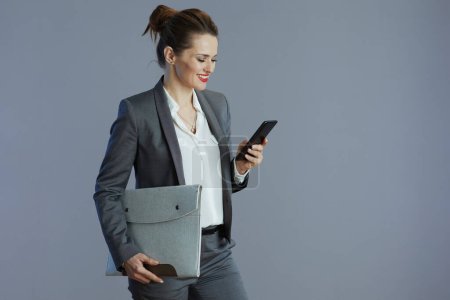 Photo for Happy stylish female employee in grey suit with smartphone and folder isolated on gray. - Royalty Free Image