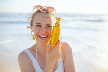 Photo for Portrait of smiling modern 40 years old woman in white swimsuit at the beach applying spf. - Royalty Free Image