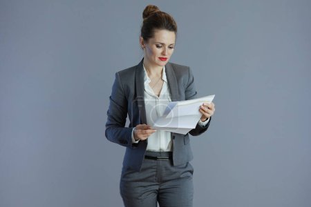 Photo for Pensive modern 40 years old woman worker in gray suit with document against grey background. - Royalty Free Image