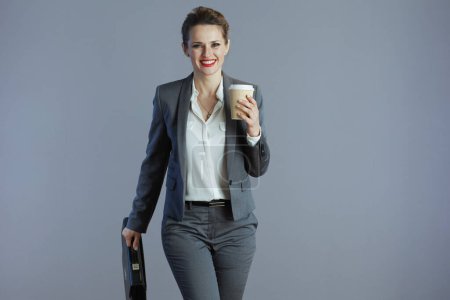Téléchargez les photos : Souriant élégant propriétaire de petite entreprise femme en costume gris avec tasse de café et mallette marchant sur fond gris. - en image libre de droit