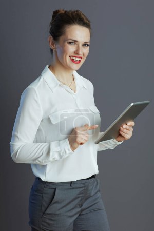 Foto de Happy stylish female employee in white blouse using applications on tablet PC on grey background. - Imagen libre de derechos