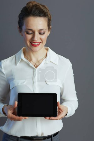 Téléchargez les photos : Smiling middle aged small business owner woman in white blouse showing tablet PC blank screen on grey. - en image libre de droit