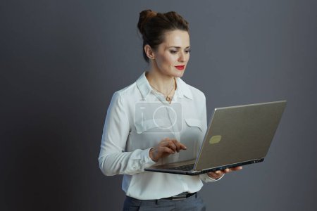 Foto de Modern 40 years old woman employee in white blouse using laptop on grey. - Imagen libre de derechos