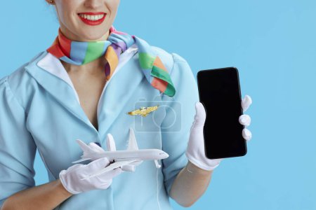 Foto de Primer plano de la mujer anfitriona del aire feliz sobre fondo azul en uniforme azul con un pequeño avión que muestra la pantalla en blanco del teléfono inteligente. - Imagen libre de derechos