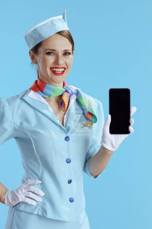 Photo for Happy elegant female flight attendant on blue background in blue uniform showing smartphone blank screen. - Royalty Free Image