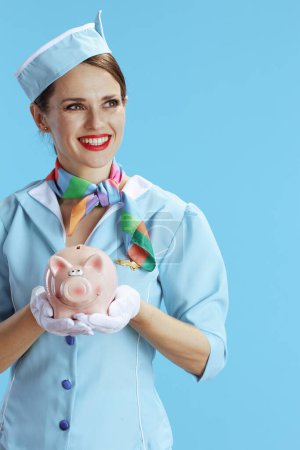Photo for Happy elegant female flight attendant on blue background in blue uniform with piggy bank. - Royalty Free Image