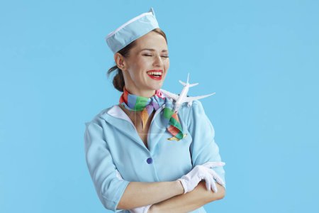 Photo for Smiling stylish female air hostess against blue background in blue uniform with a little airplane. - Royalty Free Image