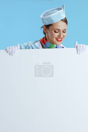 Téléchargez les photos : Souriante hôtesse de l'air féminine moderne sur fond bleu en uniforme bleu montrant tableau blanc. - en image libre de droit