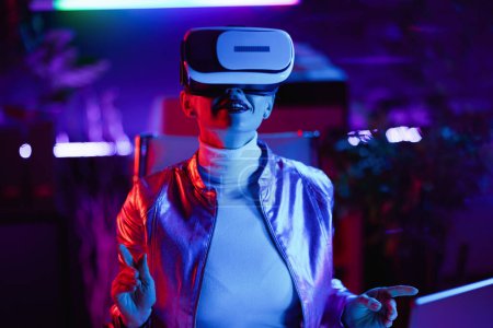Foto de Neon metaverse futuristic concept. curious stylish business woman in virtual reality goggles in modern office. - Imagen libre de derechos