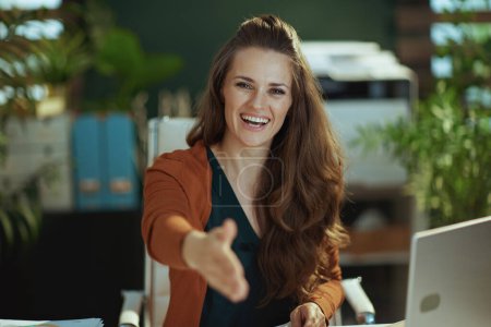 Foto de Smiling trendy 40 years old small business owner woman in the modern green office stretching hand for handshake. - Imagen libre de derechos