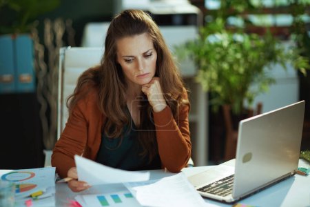 Foto de Pensive modern middle aged small business owner woman with laptop working with documents in the modern green office. - Imagen libre de derechos
