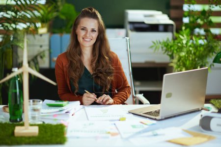 Photo for Happy 40 years old small business owner woman with laptop and wind turbine working with documents in the modern green office. - Royalty Free Image
