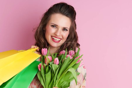 Téléchargez les photos : Portrait de femme élégante et souriante en robe florale avec bouquet de tulipes et sacs à provisions sur fond rose. - en image libre de droit