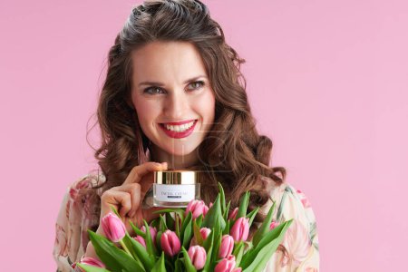Foto de Happy trendy woman with long wavy brunette hair with tulips bouquet and facial creme against pink background. - Imagen libre de derechos