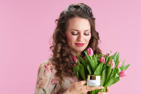 Photo for Relaxed stylish middle aged woman in floral dress with tulips bouquet and cosmetic jar against pink background. - Royalty Free Image
