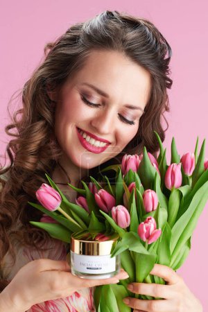 Foto de Happy elegant female with long wavy brunette hair with tulips bouquet and cosmetic jar against pink background. - Imagen libre de derechos