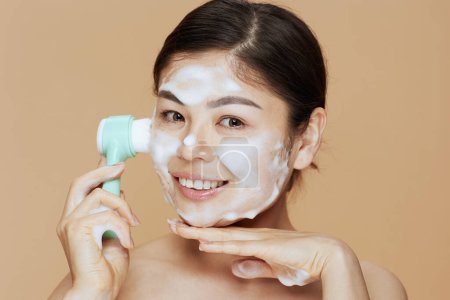 Foto de Retrato de mujer asiática joven con masajeador lavando la cara sobre fondo beige. - Imagen libre de derechos