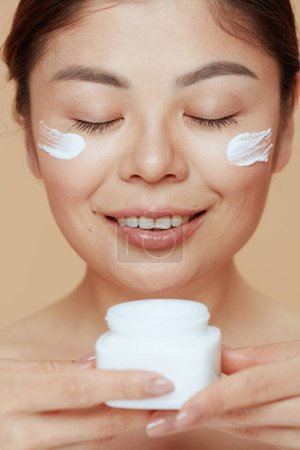 Téléchargez les photos : Modern woman with facial cream jar and facial cream on face on beige background. - en image libre de droit