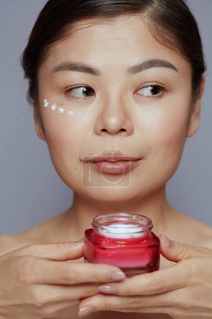 Photo for Young asian female with facial cream jar and eye cream on face against blue background. - Royalty Free Image
