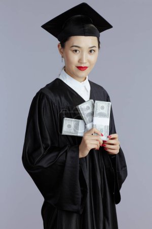 Téléchargez les photos : Happy modern female asian graduate student with money isolated on gray background. - en image libre de droit
