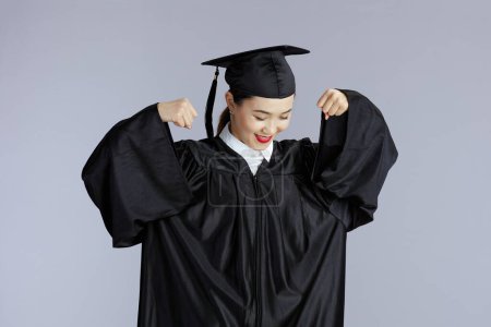 Foto de Proud young female asian graduate student in graduation gown with cap showing biceps isolated on gray. - Imagen libre de derechos