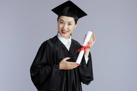 Foto de Happy young graduate student asian woman with diploma against grey background. - Imagen libre de derechos