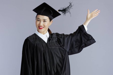 Téléchargez les photos : Joyeux moderne diplômé asiatique femme en robe de graduation avec casquette lancer glands isolé sur fond gris. - en image libre de droit