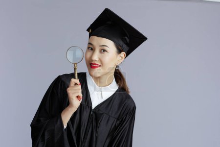 Photo for Modern female asian graduate student with magnifying glass against grey background. - Royalty Free Image