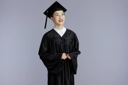 Téléchargez les photos : Smiling young graduate student asian woman in graduation gown with cap looking into the distance isolated on gray. - en image libre de droit