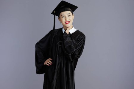 Foto de Feliz joven estudiante de posgrado mujer asiática en vestido de graduación con gorra mirando espacio de copia contra fondo gris. - Imagen libre de derechos