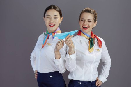 Photo for Smiling elegant air hostess women in blue skirt, white shirt and scarf with paper airplane isolated on grey. - Royalty Free Image