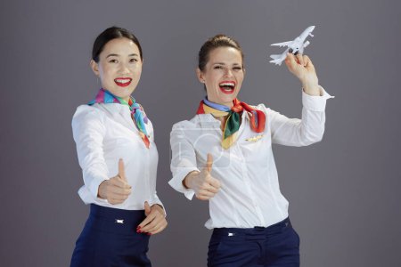 Téléchargez les photos : Joyeuses hôtesses de l'air féminines élégantes en jupe bleue, chemise blanche et écharpe avec un petit avion isolé sur fond gris. - en image libre de droit