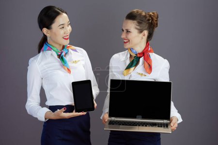 Photo for Happy elegant stewardess women in blue skirt, white shirt and scarf showing digital tablet blank screen and and laptop blank screen isolated on grey. - Royalty Free Image
