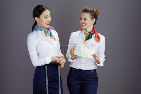 Foto de Happy elegant female air hostesses in blue skirt, white shirt and scarf with flight tickets and travel bag against gray background. - Imagen libre de derechos