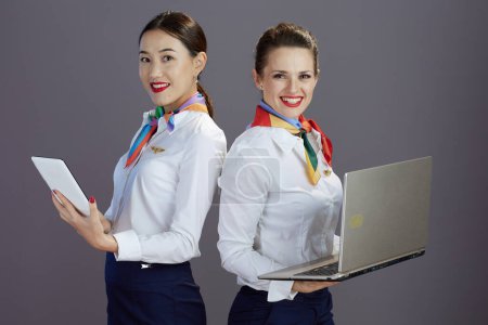 Photo for Happy elegant stewardess women in blue skirt, white shirt and scarf with laptop using tablet PC isolated on gray. - Royalty Free Image