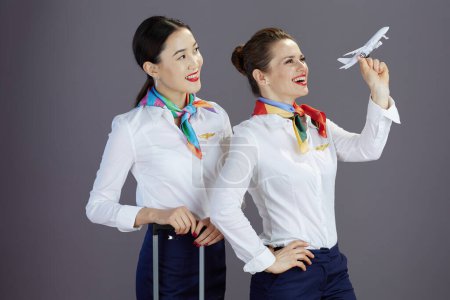 Foto de Happy modern female flight attendants in blue skirt, white shirt and scarf with a little airplane and trolley bag isolated on grey background. - Imagen libre de derechos
