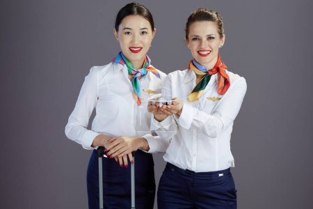 Foto de Smiling stylish female stewardesses in blue skirt, white shirt and scarf with a little airplane and trolley bag isolated on grey background. - Imagen libre de derechos