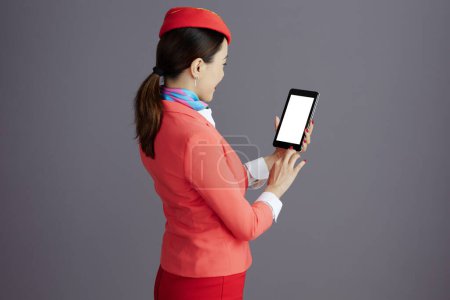 Photo for Seen from behind modern flight attendant asian woman in red skirt, jacket and hat uniform with tablet PC isolated on gray. - Royalty Free Image