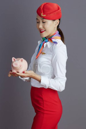 Photo for Happy elegant asian female flight attendant in red skirt and hat uniform with piggy bank isolated on gray background. - Royalty Free Image