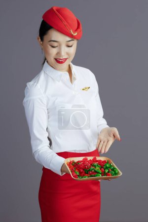 Téléchargez les photos : Heureux moderne air hôtesse asiatique femme en rouge jupe et chapeau uniforme avec des bonbons isolés sur gris. - en image libre de droit