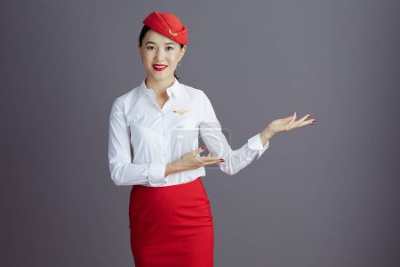 Foto de Smiling elegant air hostess asian woman in red skirt and hat uniform welcoming isolated on grey background. - Imagen libre de derechos