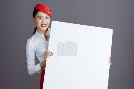 Foto de Happy modern asian female stewardess in red skirt and hat uniform with blank billboard isolated on gray. - Imagen libre de derechos