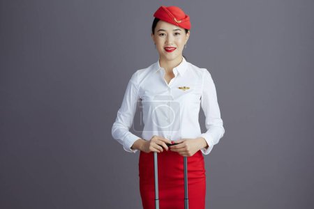 Foto de Feliz moderno aire azafata asiático mujer en rojo falda y sombrero uniforme con rueda bolsa contra gris fondo. - Imagen libre de derechos