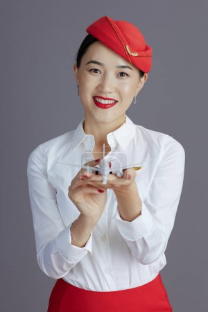 Photo for Happy elegant asian female stewardess in red skirt and hat uniform with a little airplane against grey background. - Royalty Free Image
