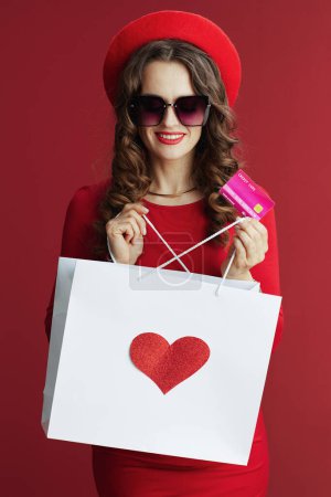 Photo for Happy Valentine. smiling stylish female with long wavy hair in red dress and beret with credit card and shopping bag and heart isolated on red. - Royalty Free Image