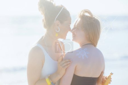Téléchargez les photos : Mère élégante heureuse et fille adolescente à la plage en vêtements de plage appliquant de la crème solaire. - en image libre de droit