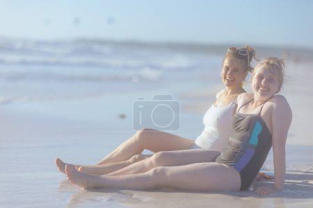 Téléchargez les photos : Souriant élégant mère et enfant en maillot de bain assis à la plage. - en image libre de droit