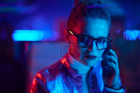 Foto de Concepto futurista neón metaverso. Retrato de mujer moderna pensativa hablando en un teléfono inteligente en la oficina moderna. - Imagen libre de derechos