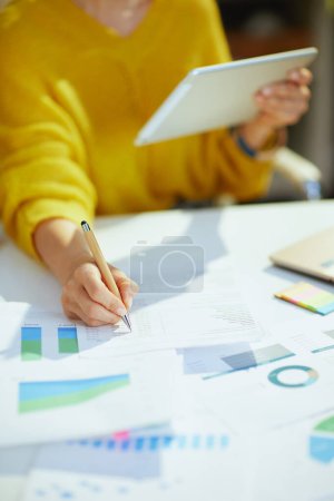 Photo for Closeup on middle aged small business owner woman in yellow sweater using tablet PC and working with documents in the green office. - Royalty Free Image