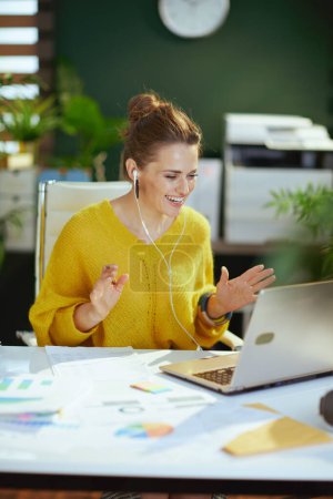 Foto de Feliz moderno dueño de la pequeña empresa mujer en suéter amarillo con el ordenador portátil que tiene la web cam reunión en la moderna oficina verde. - Imagen libre de derechos